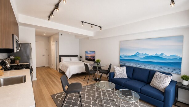 A modern hotel room with a kitchenette, a bed, a sofa, a dining area, and a large window with a mountain view. - Perfect Studio Downtown Sidney (Victoria)