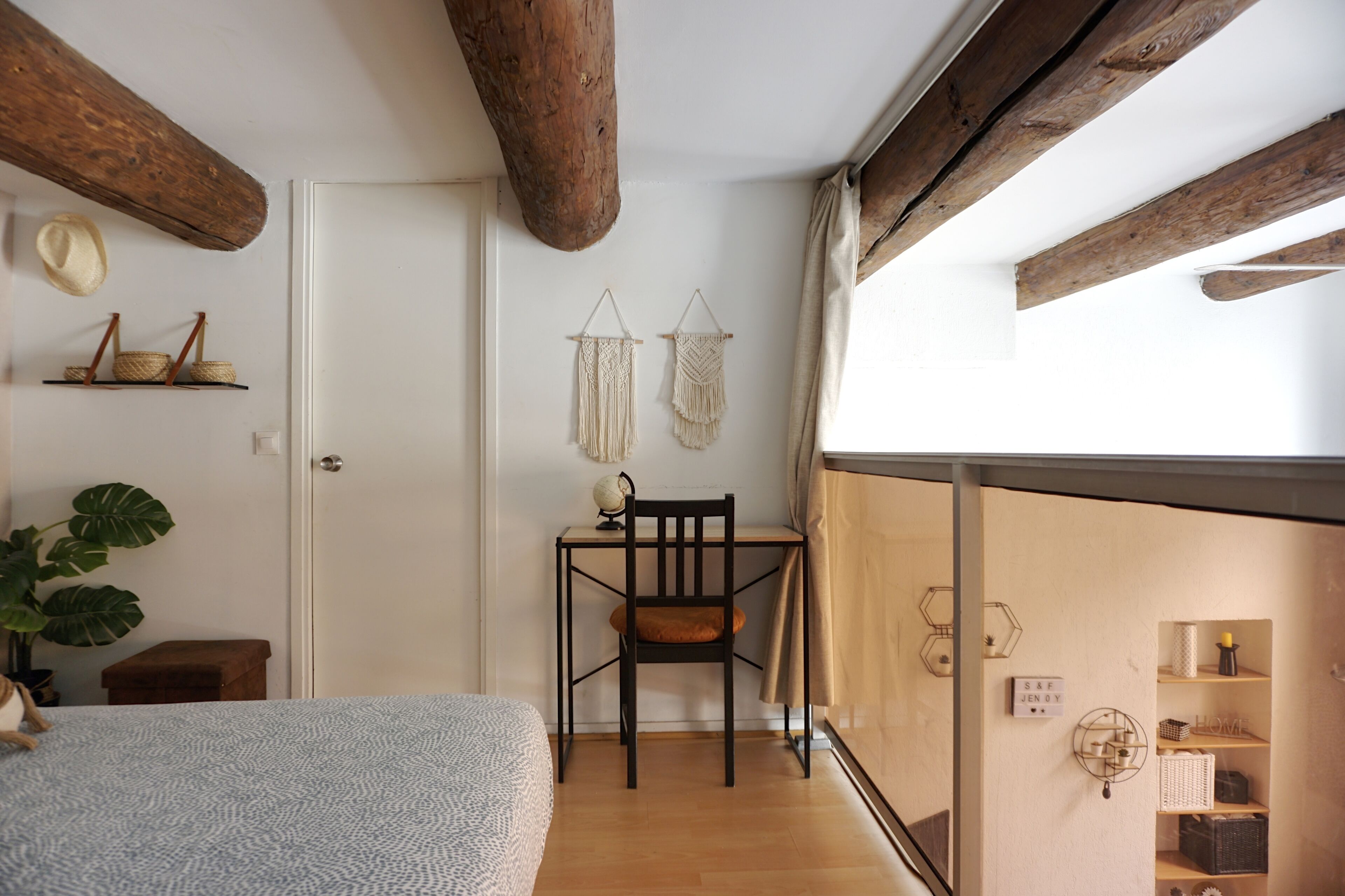 A room with a bed, a desk with a chair, wooden beams, a plant, and a view through a glass partition.