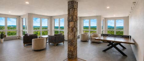 A spacious room with a ping pong table, multiple seating areas, and large windows offering a view of greenery.