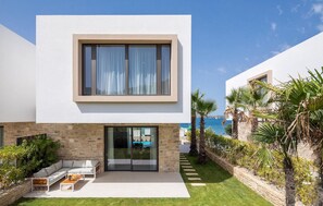 Una casa moderna de dos plantas con fachada de piedra, puerta de cristal de entrada, un patio con mobiliario exterior y vista al mar.