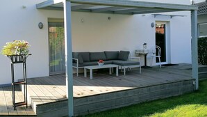 Un espace extérieur aménagé avec un coin salon couvert, comprenant un canapé et une table, une plante en pot et une petite lampe de table.