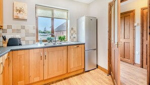 A modern kitchen with wooden cabinets, a stainless steel refrigerator, and a window with a view of a house. - Lake View 2 - UK45578 (Louth)