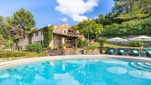 Un área de piscina con un edificio de piedra, asientos al aire libre y abundante vegetación.