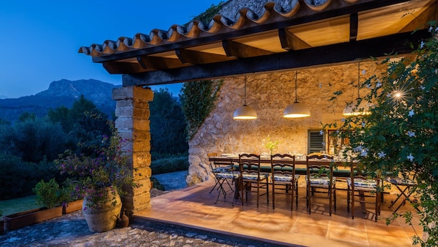 Un espacio de comedor al aire libre con paredes de piedra y mobiliario de madera, rodeado de follaje y un paisaje montañoso.
