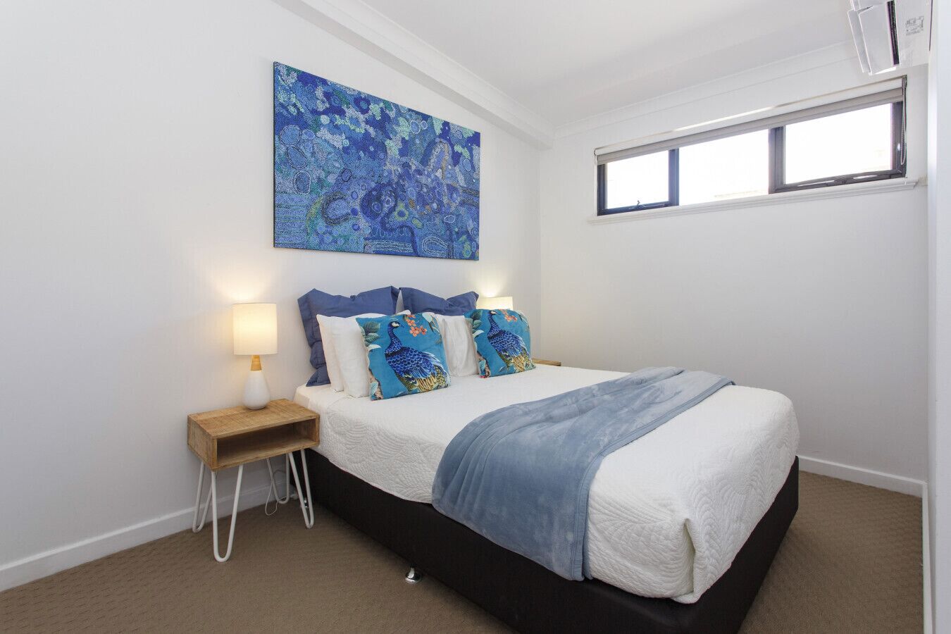 A bedroom with a bed, a nightstand, a lamp, a window, and a wall-mounted painting.