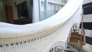 A hammock suspended between wooden chairs in a room with tiled flooring and a television in the background. - Prainha PÉ NA Praia (Prainha Arraial do Cabo)