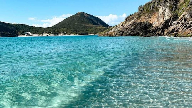 Clear turquoise water with visible underwater patterns, a mountainous landscape, and rocky terrain. - Prainha PÉ NA Praia (Prainha Arraial do Cabo)