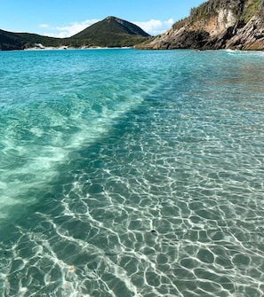 A clear turquoise sea with visible underwater patterns, a rocky coastline, and a mountain in the distance. - Prainha PÉ NA Praia (Prainha Arraial do Cabo)