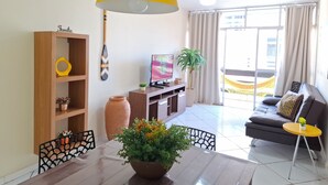 A living room with a wooden bookshelf, a TV, a potted plant, a sofa, and a dining table with chairs. - Prainha PÉ NA Praia (Prainha Arraial do Cabo)