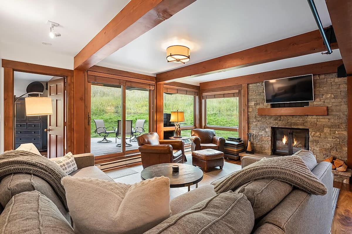 A cozy living room with a fireplace, leather furniture, and large windows offering a view of the outdoors.