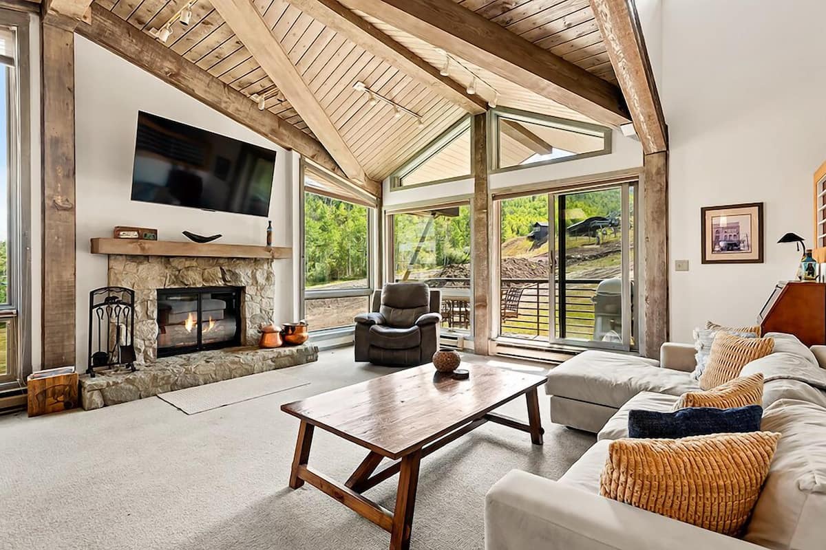 A spacious living room with a stone fireplace, a flat-screen TV mounted above it, and a large window offering a view of the outdoors.