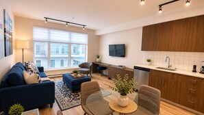 A modern living room with a sofa, coffee table, and chairs, a kitchen with wooden cabinets and a sink, and a large window providing natural light. - Perfect Brand New Suite In The Heart of Sidney (Sidney)