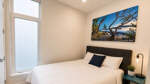 A bedroom with a bed, a window, a wall-mounted painting, a bedside table with a lamp, and a potted plant. - Perfect Brand New Suite In The Heart of Sidney (Sidney)