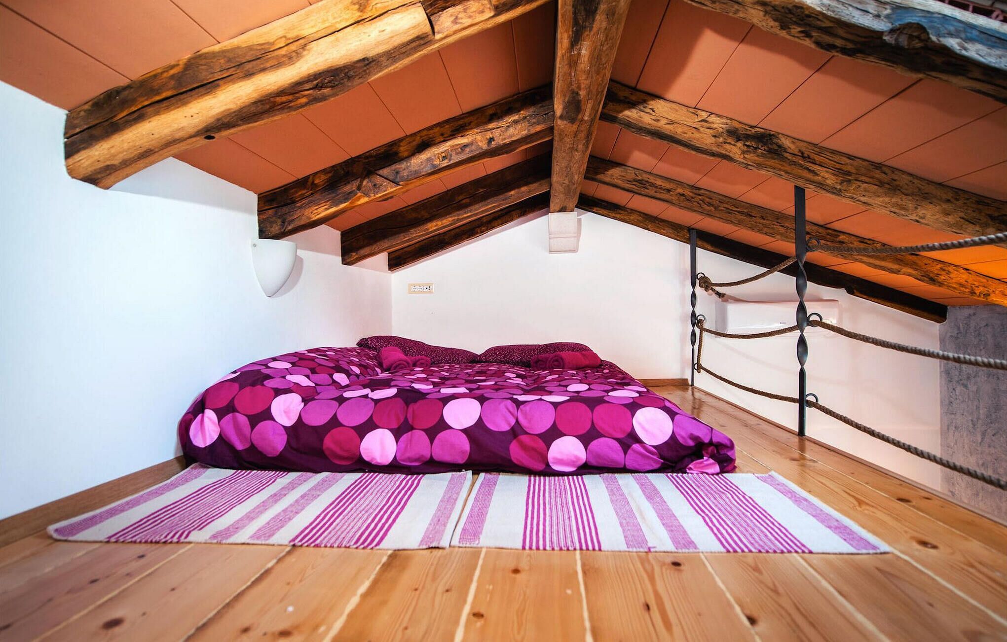 Une chambre avec un plafond en pente, des poutres en bois, un lit recouvert d’une couvre-lit à motifs, un tapis à rayures et un luminaire fixé au mur.