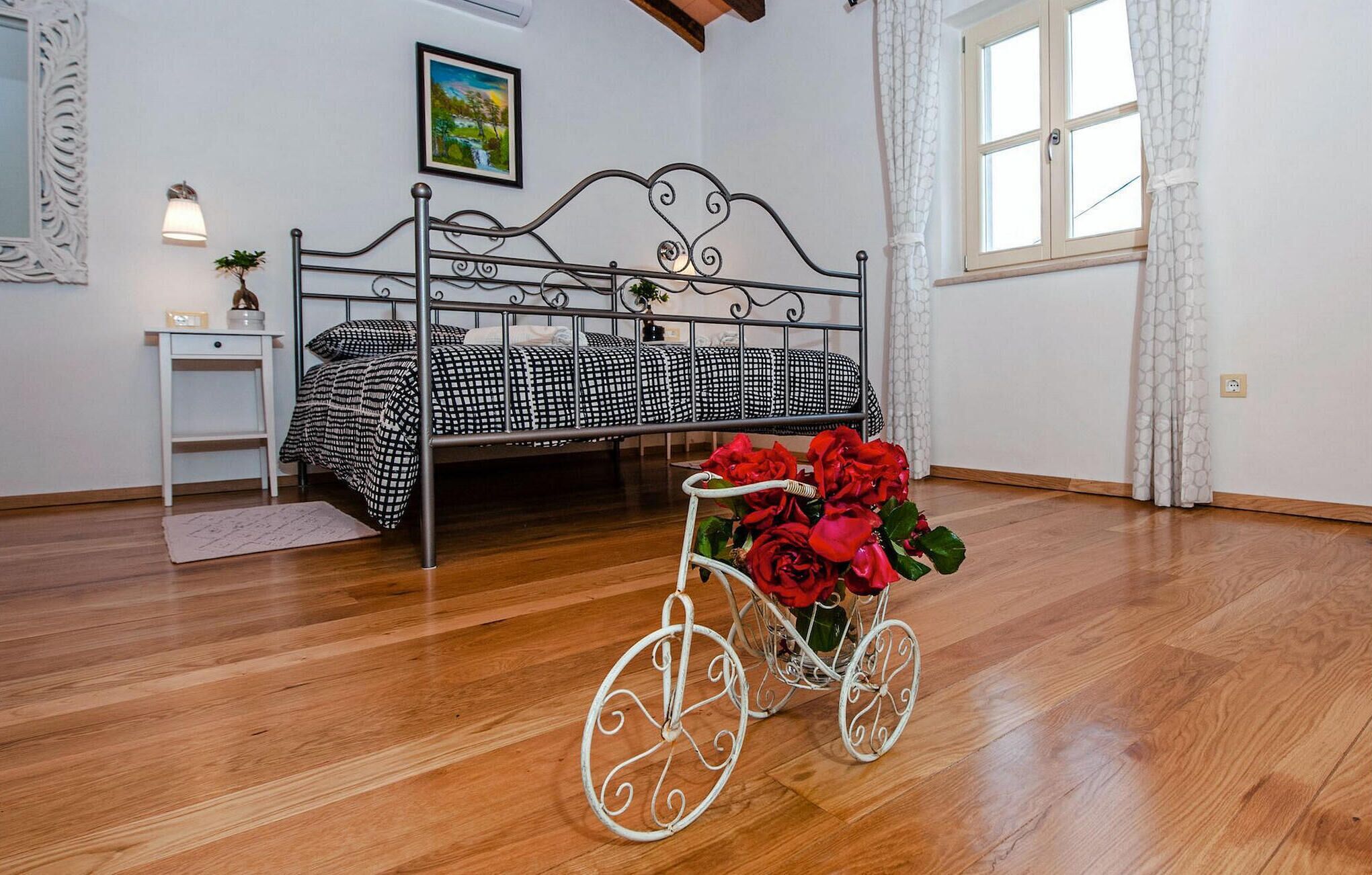 Et soveværelse med en sengegavl i metal, et natbord, en vindue med gardiner og en cykel pyntet med blomster.