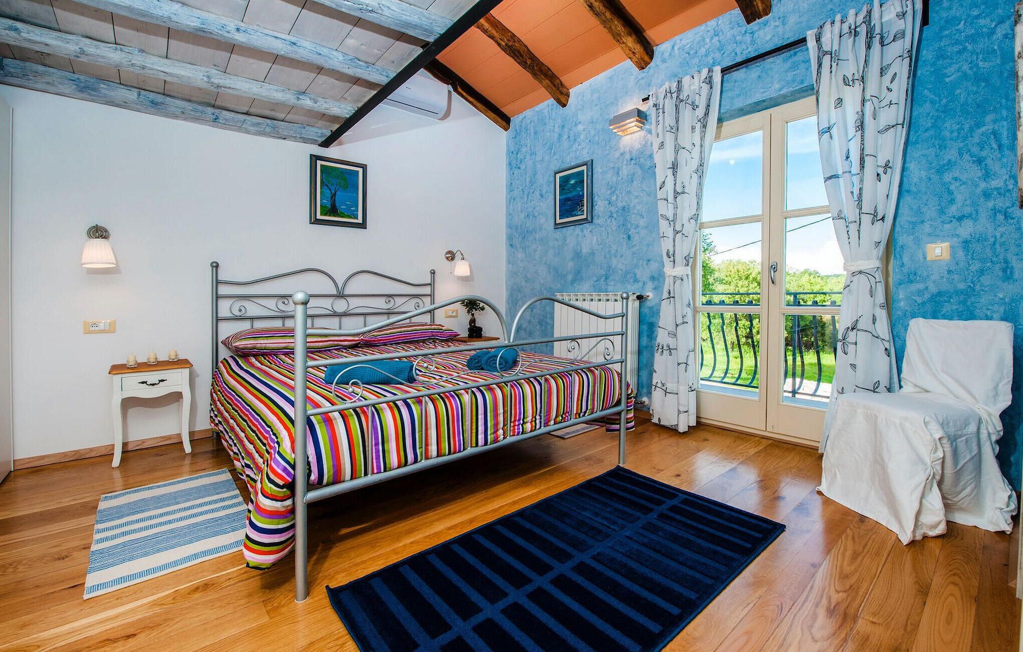 A bedroom with a metal bed frame, a small bedside table, a window with curtains, and a view of greenery outside.
