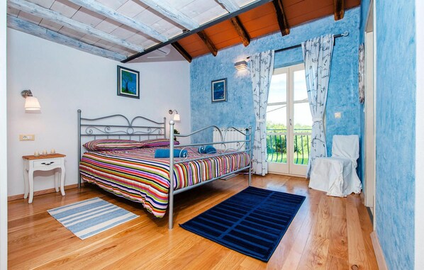 A bedroom with a metal bed frame, a striped bedspread, a small bedside table, a nightstand, a window with curtains, and a view of greenery outside.