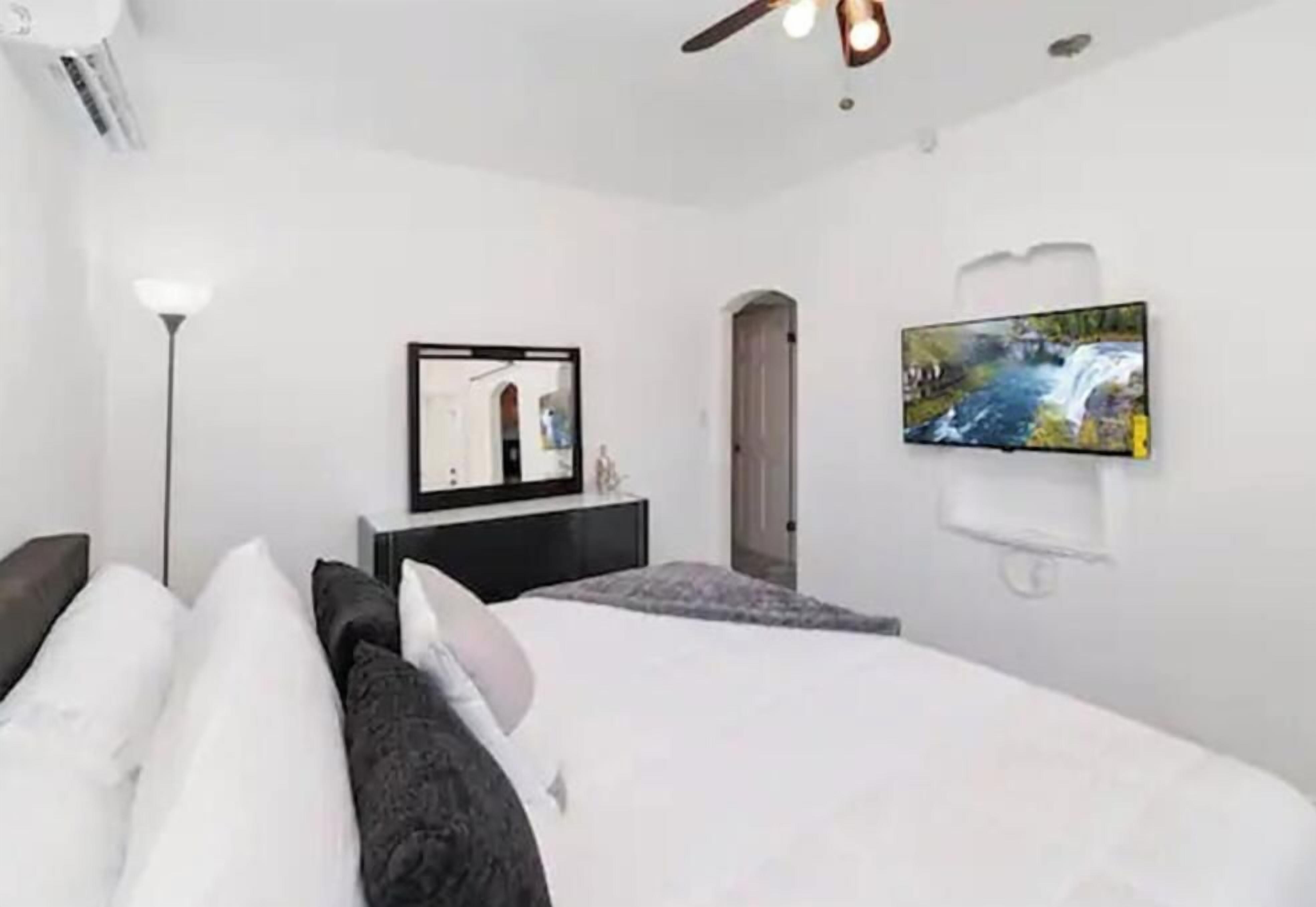A bedroom with a bed, a flat-screen TV mounted on the wall, a bedside table, and a door leading to another room.