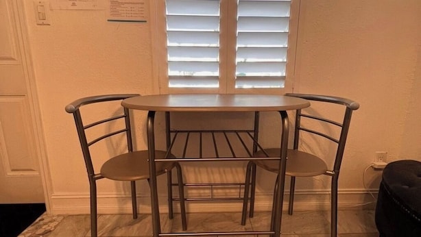 Un pequeño comedor con una mesa redonda y cuatro sillas, una ventana con persianas y una vista parcial de una pared con un interruptor de luz y un papel.