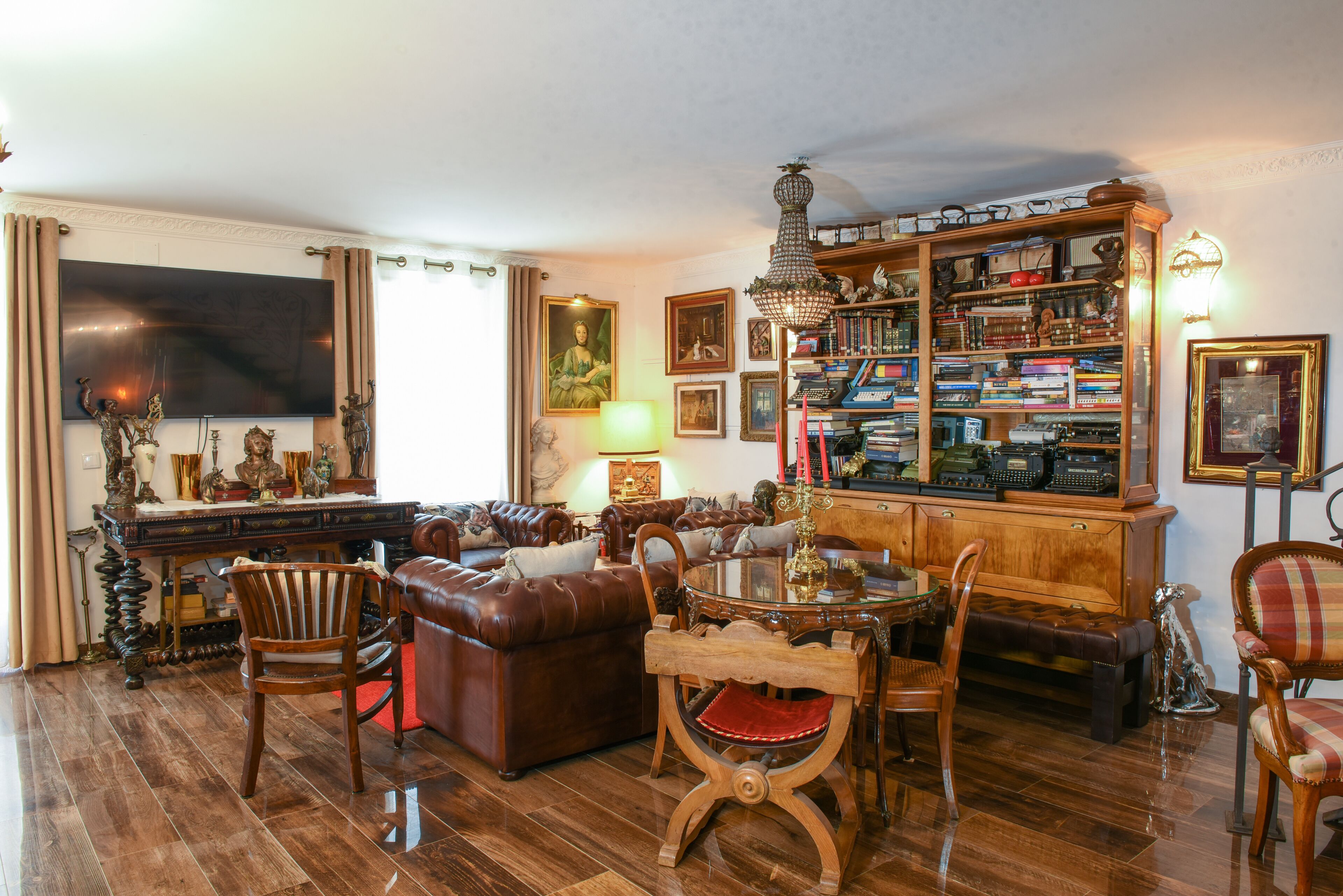 A living room with a brown leather sofa, wooden chairs, a glass coffee table, a bookshelf, and a flat-screen TV.