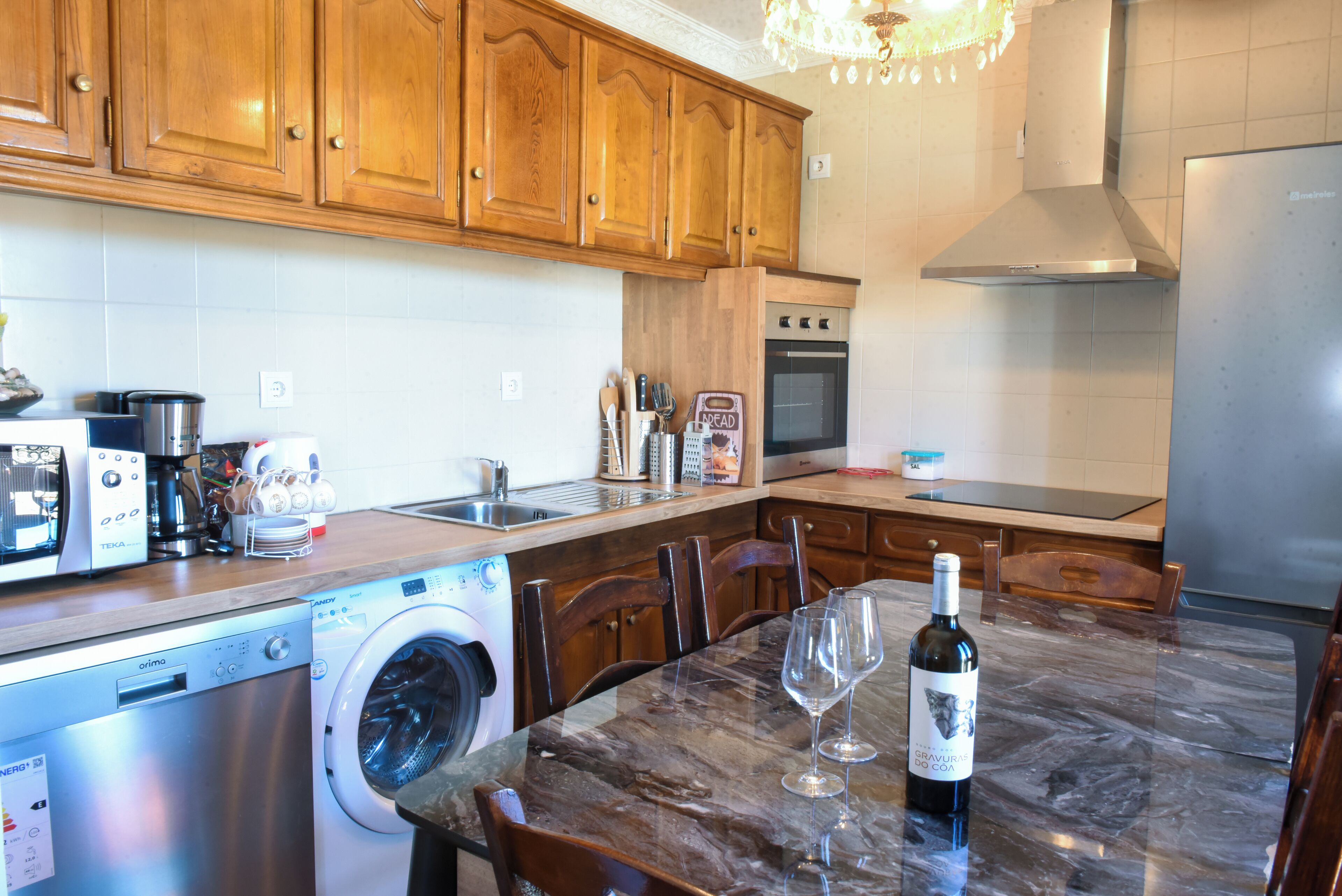 A kitchen with wooden cabinets, a stainless steel oven, a washing machine, and a marble dining table with a wine bottle and glass.