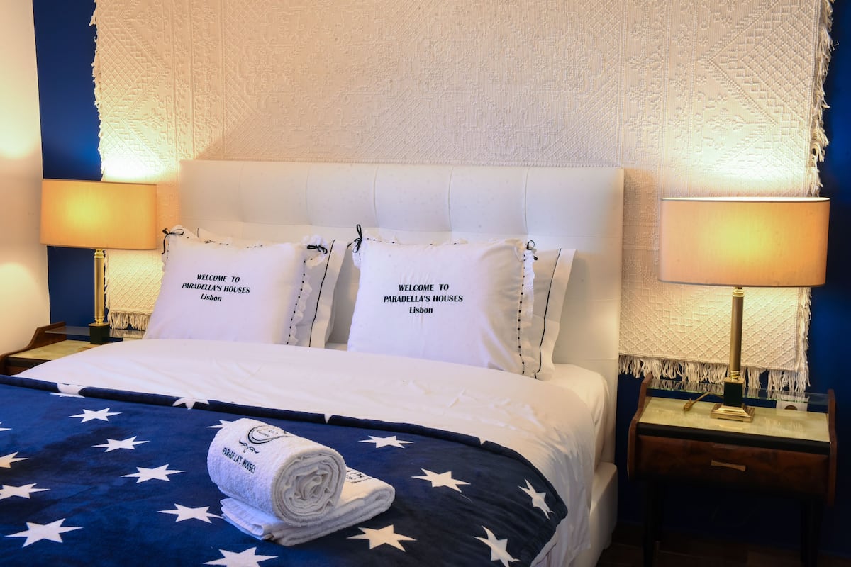 A hotel room with a bed featuring a blue and white star-patterned bedspread, two pillows with "Welcome to Paradella's Houses Lisbon" text, and a rolled towel on the bed.