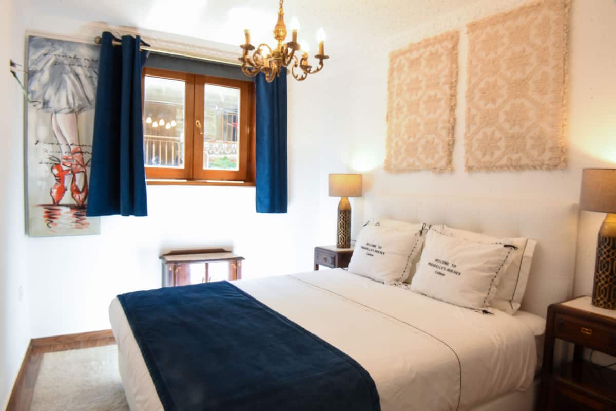 A neatly arranged bedroom with a bed, a nightstand, a lamp, a window with curtains, and a chandelier.