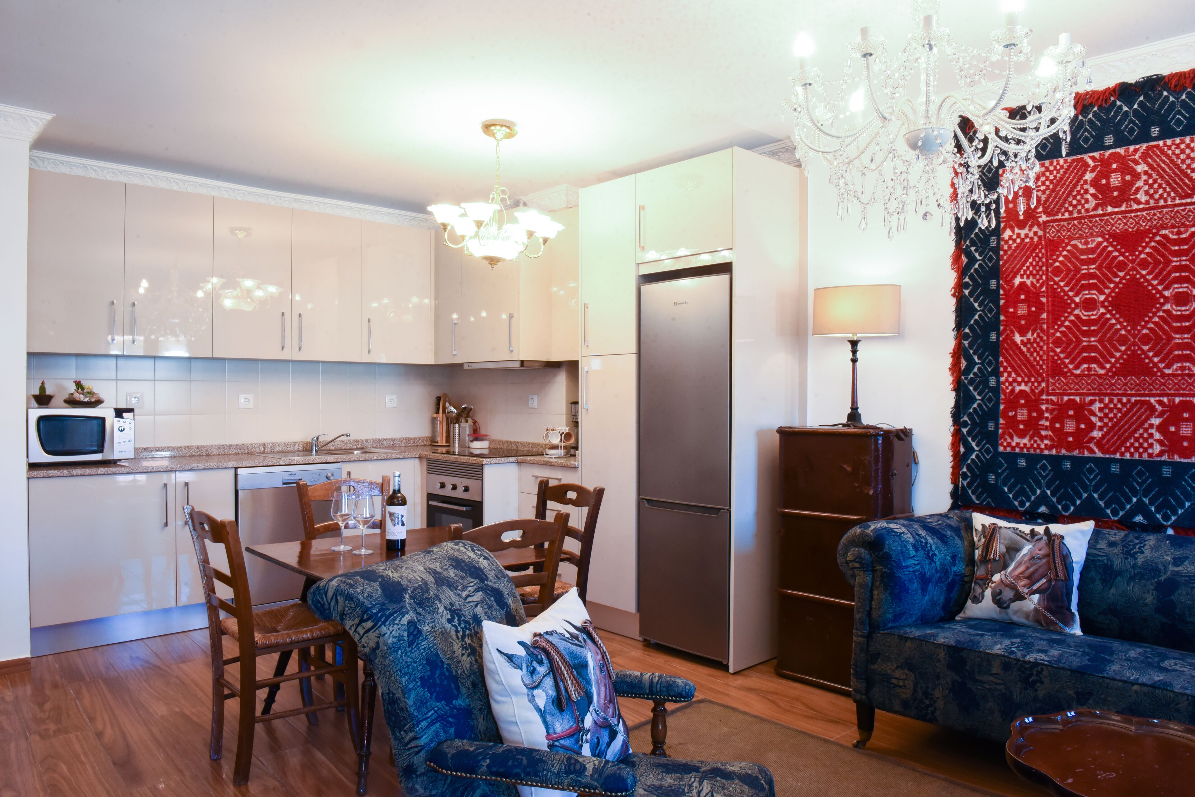 Una cocina moderna con armarios blancos, un refrigerador de acero inoxidable y un comedor con un sofá tapizado en azul y sillas de madera.