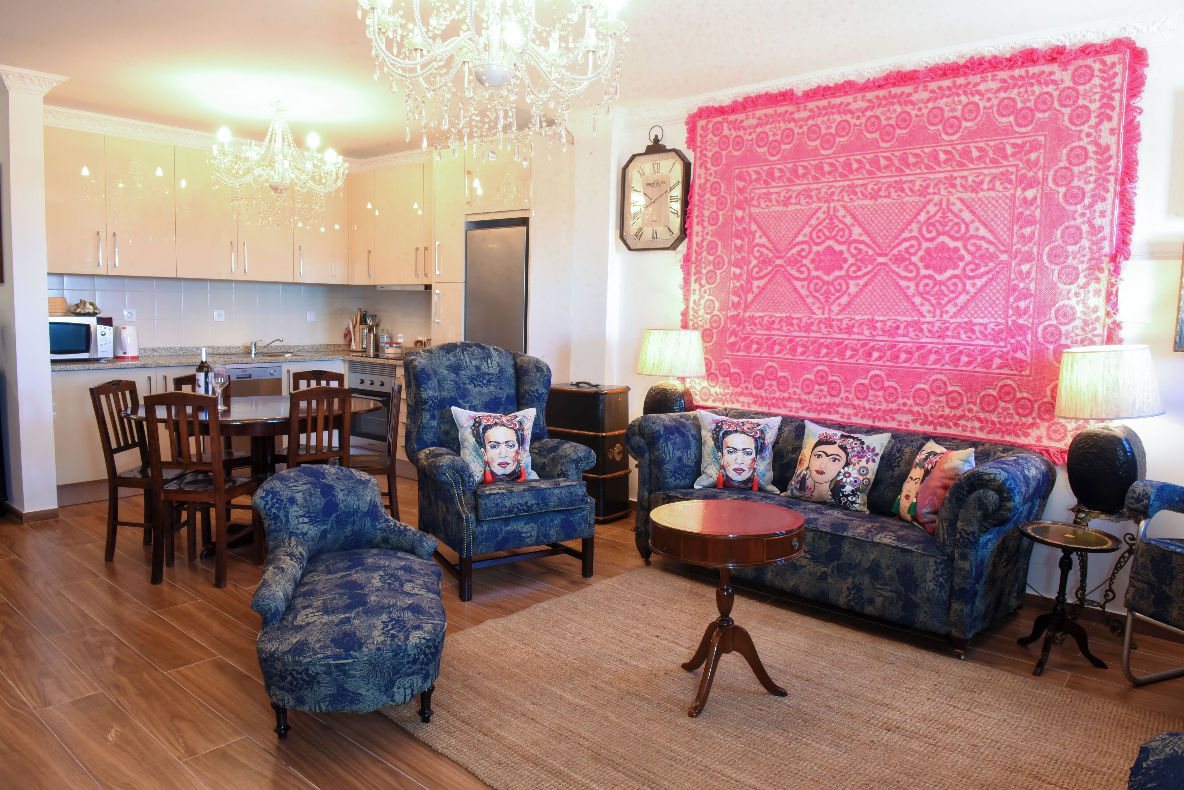 Una sala de estar con un sofá azul, un comedor con sillas de madera, una cocina con gabinetes blancos y un tapiz rosa grande en la pared.
