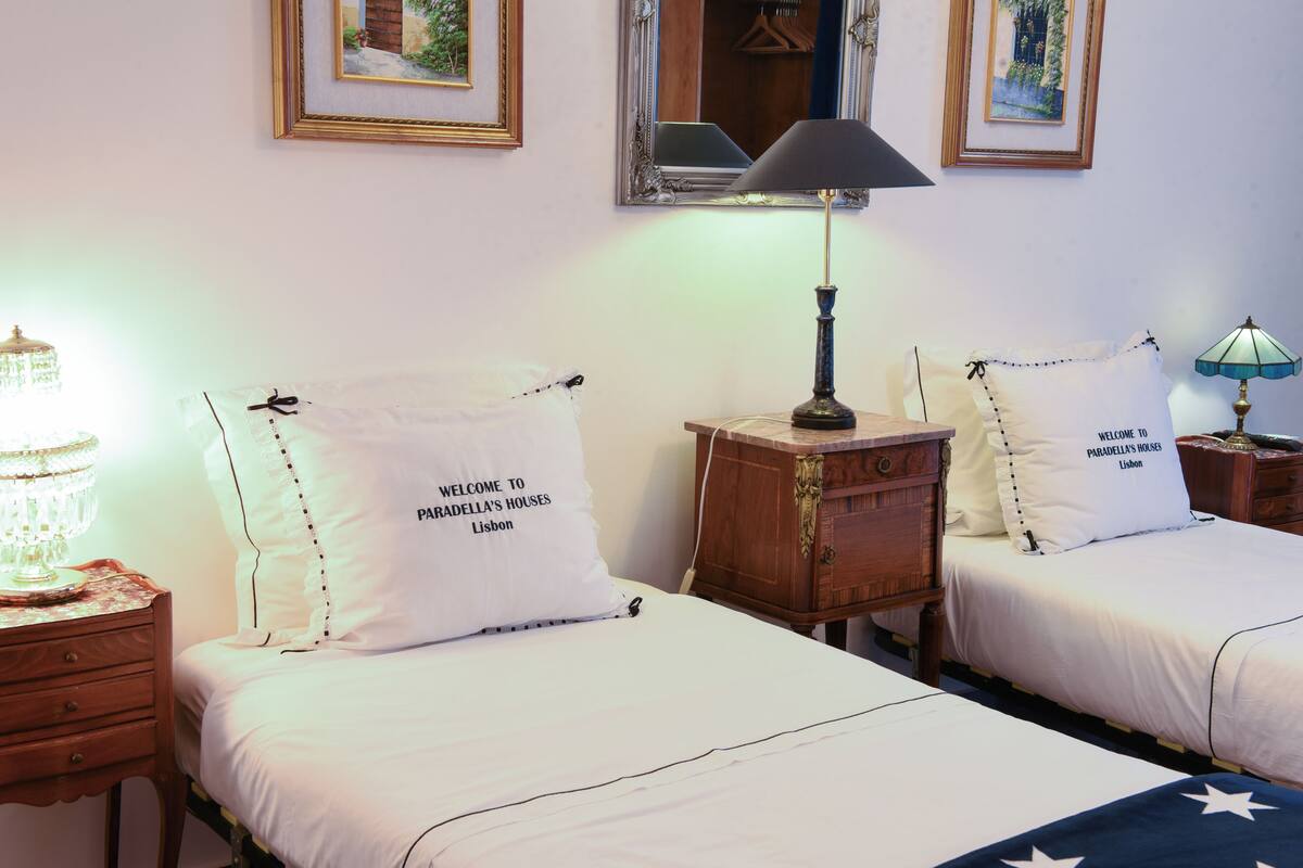 A hotel room with two beds, each with a pillow stating "Welcome to Paradella's Houses Lisbon." There is a lamp on each nightstand, and a chandelier on the left side.