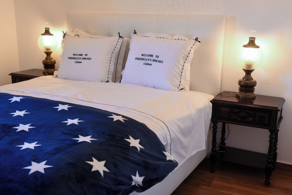 A bed with a blue blanket featuring white stars, two pillows with "Welcome to Paradella's Houses Lisbon" text, and two bedside lamps on wooden nightstands.