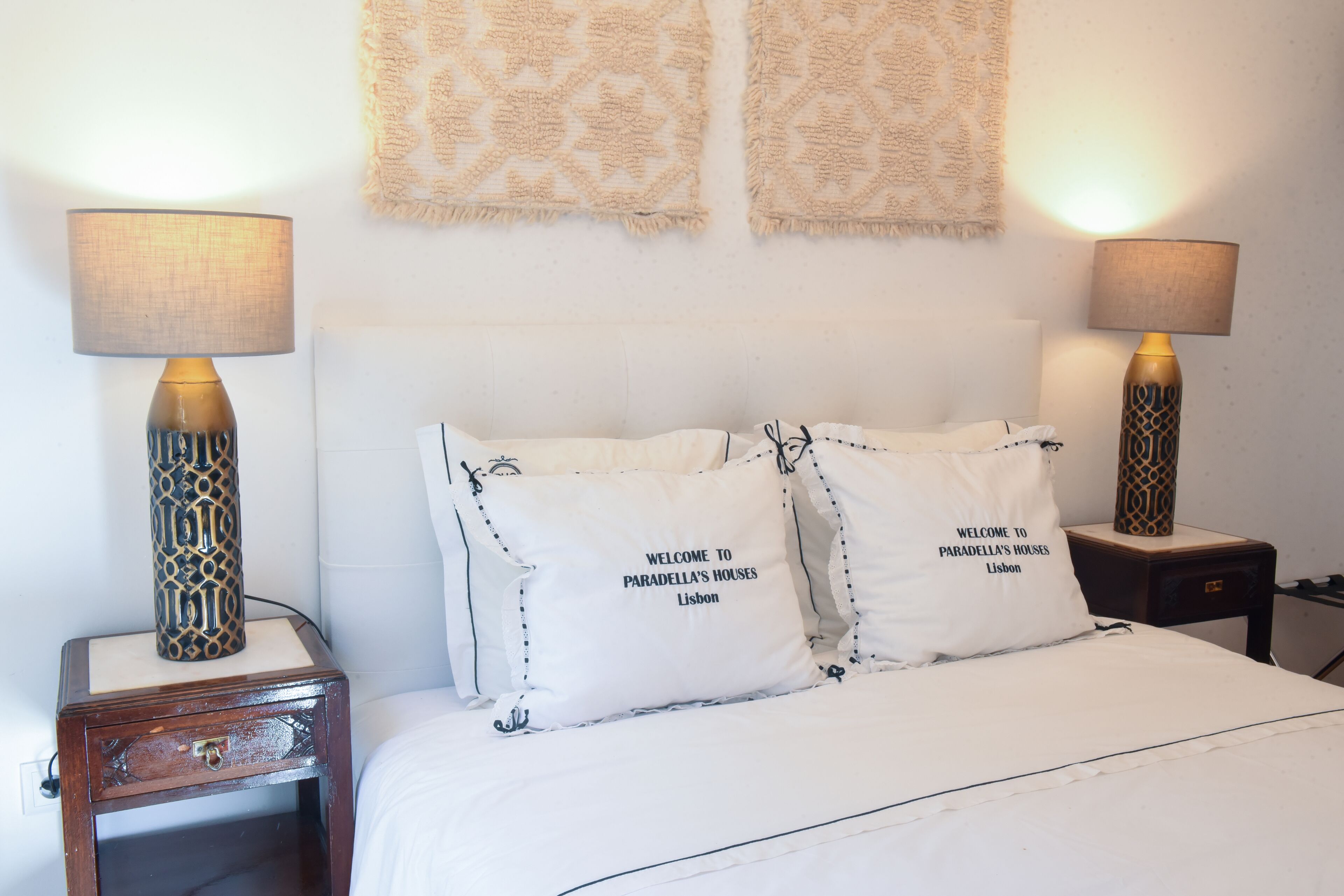 A neatly made bed with two pillows, each with "Welcome to Paradella's Houses Lisbon" text, flanked by two bedside lamps with patterned bases.