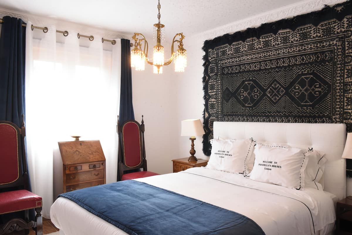 A bedroom with a large bed, a wooden nightstand, a red chair, a wooden dresser, and a patterned tapestry on the wall.