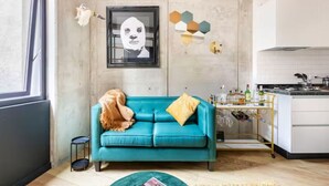 Un salon moderne avec un canapé de couleur turquoise, une petite table ronde et un coin cuisine doté d’un meuble à bouteilles.