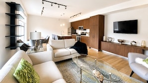 Ein modernes Wohnzimmer mit Sofa, Couchtisch und Fernseher. Es gibt eine Küchenzeile mit Schränken und Geräten.