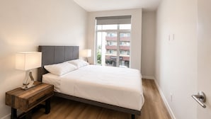 Ein Schlafzimmer mit einem Bett, zwei Nachttischlampen, einem Holfnachtkästchen und Blick auf ein Gebäude durch ein Fenster.