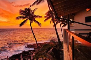 Vista del atardecer desde un balcón con palmeras y una playa.