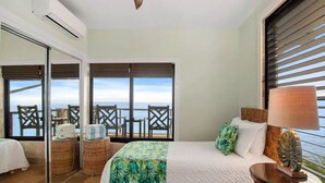 Quarto de hotel com uma cama, mesa de cabeceira, abajur e vista para o oceano através de grandes janelas.