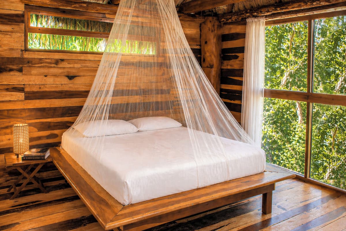 Un lit en bois surmonté d’un baldaquin, une table de chevet avec une lampe et une fenêtre donnant sur un paysage verdoyant.
