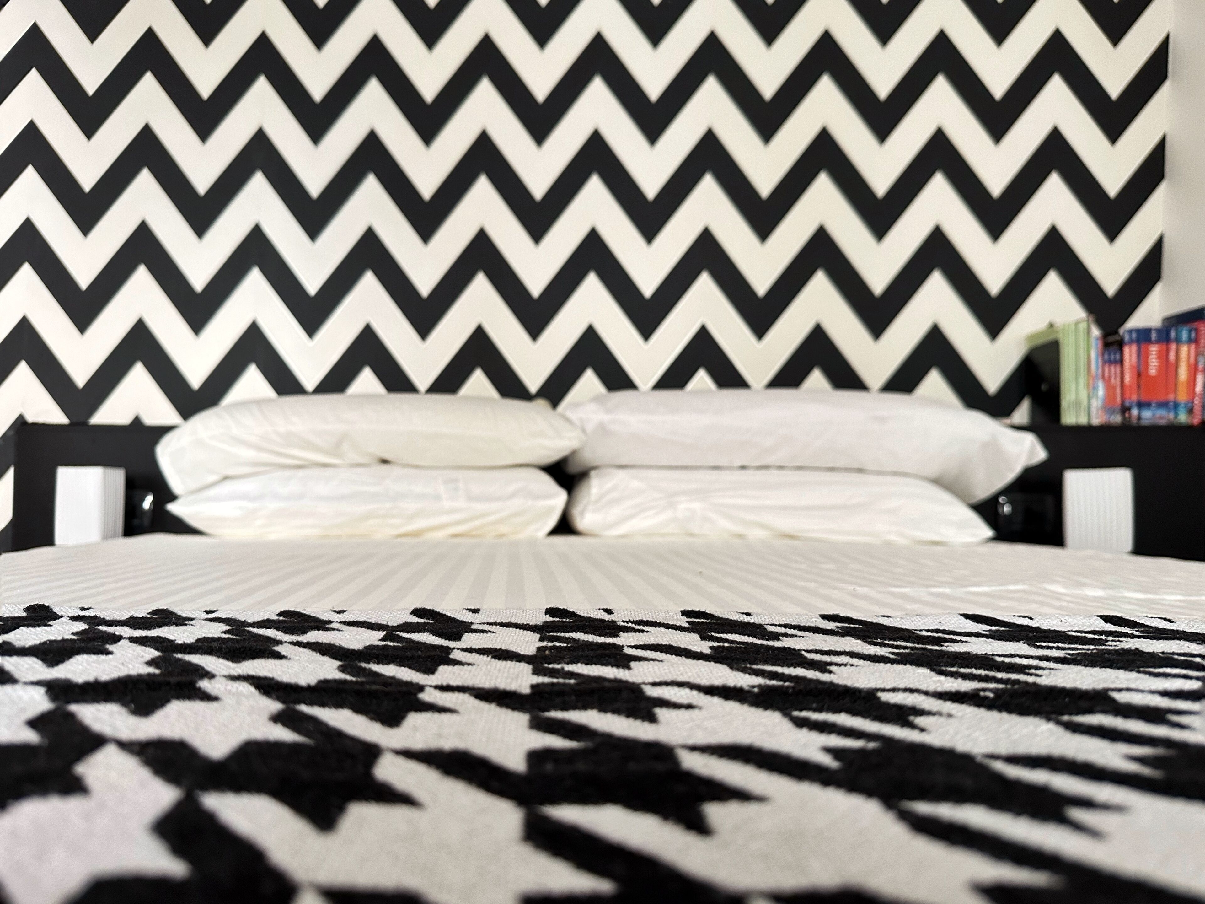 Una cama con una colcha estampada, un diseño geométrico en blanco y negro en la pared y una estantería con libros.