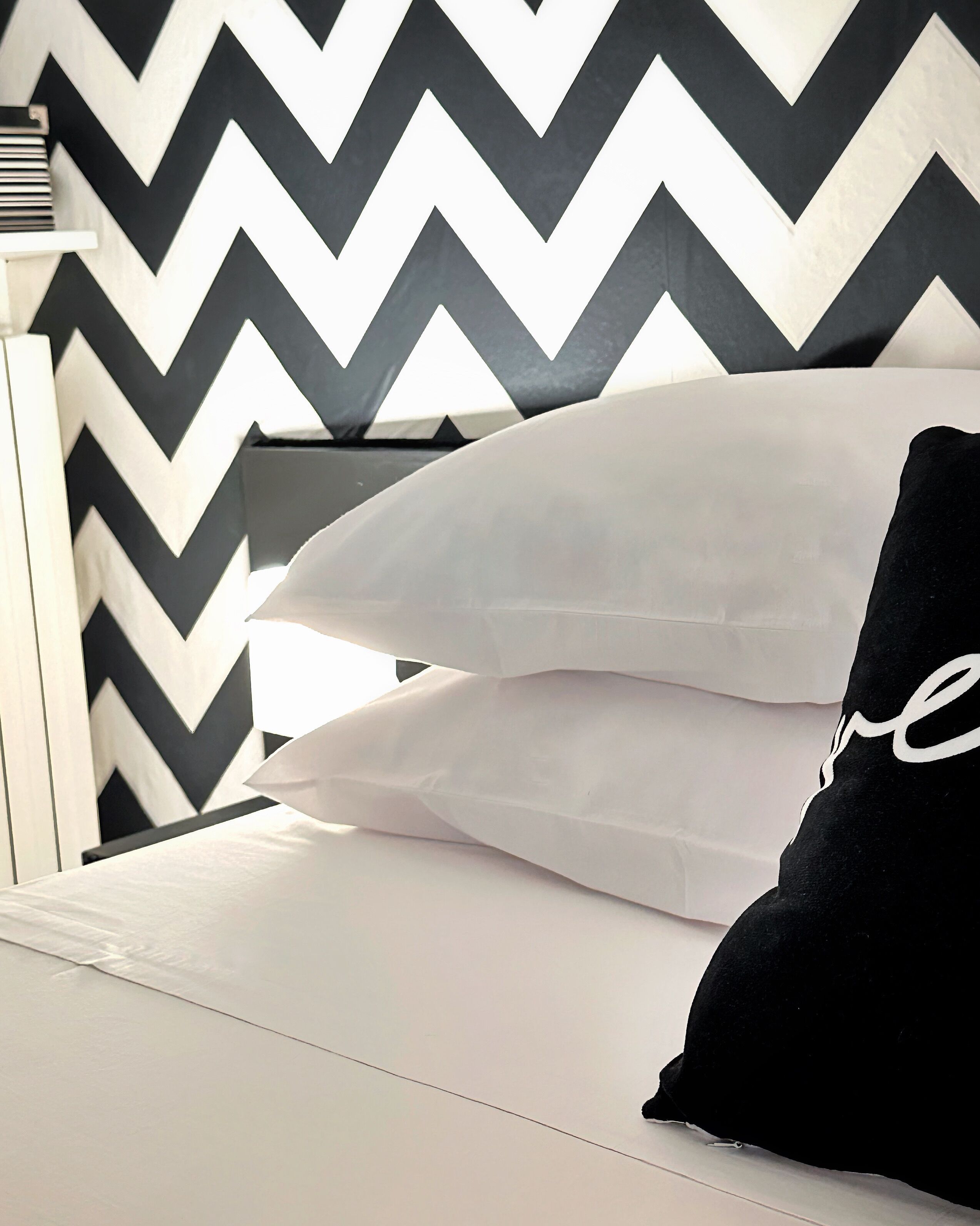 A bed with white bedding and two pillows, one white and one black, against a wall with a black and white chevron pattern.