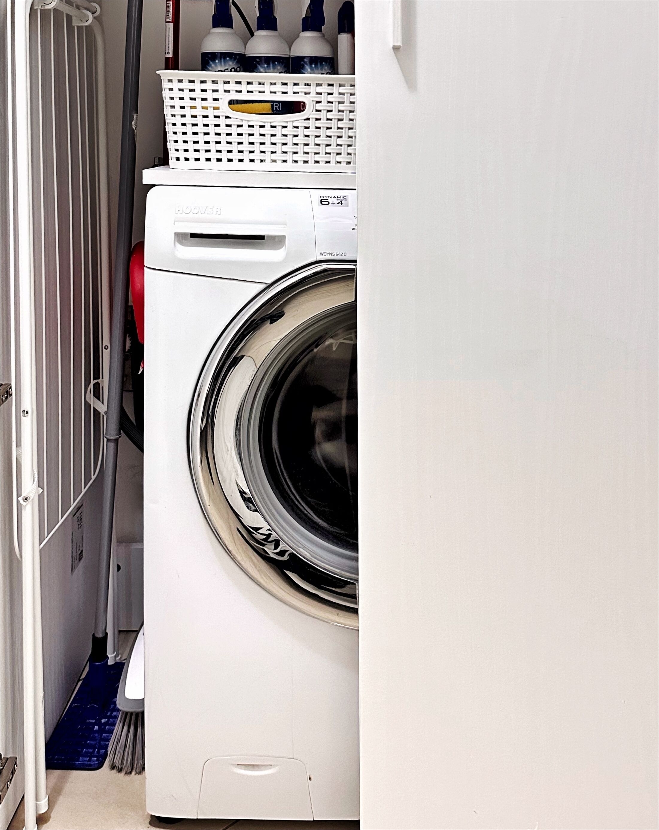 A front-loading washing machine in a laundry room with a basket on top and a mop leaning against the wall.