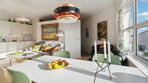 Una cocina moderna con armarios blancos, un comedor con mesa blanca y sillas verdes, y un amplio ventanal con vista al cielo.