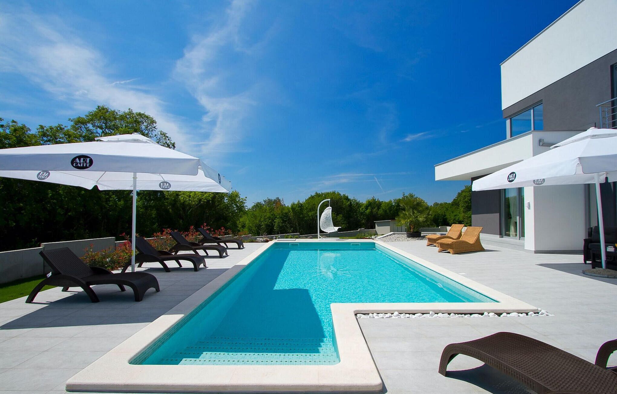 Un espace piscine extérieur moderne, doté de chaises longues et de parasols, entouré de verdure et surplombé par un ciel dégagé.