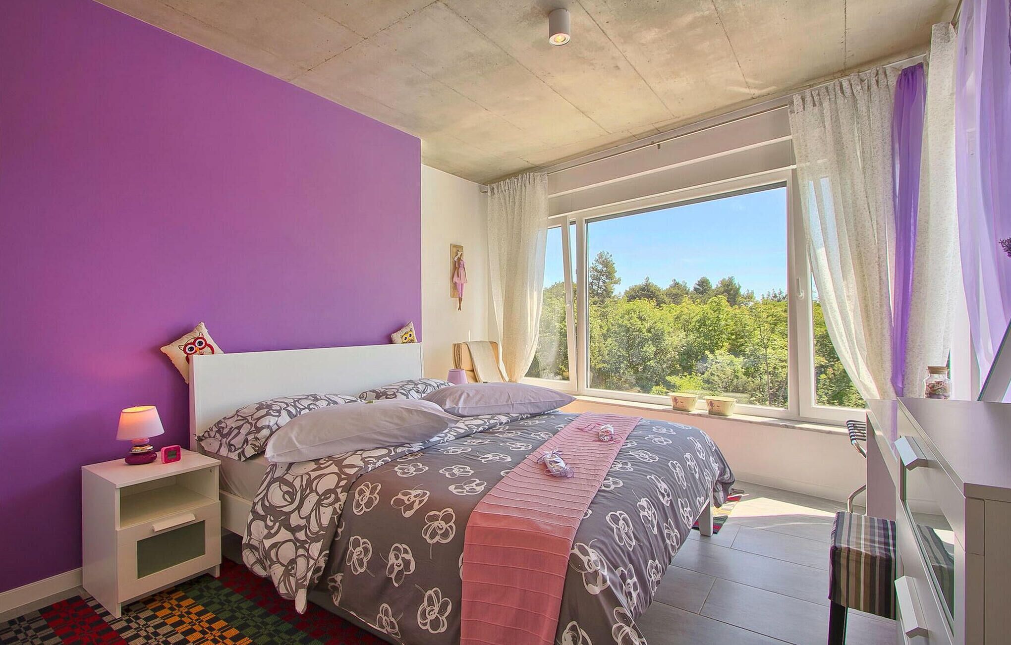 Une chambre avec un grand lit, une table de chevet, une fenêtre avec des rideaux et une vue sur les arbres.