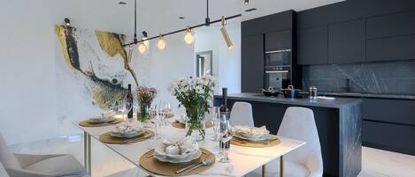 A modern dining area with a white table set for four, gold accents, and a contemporary kitchen with dark cabinetry.