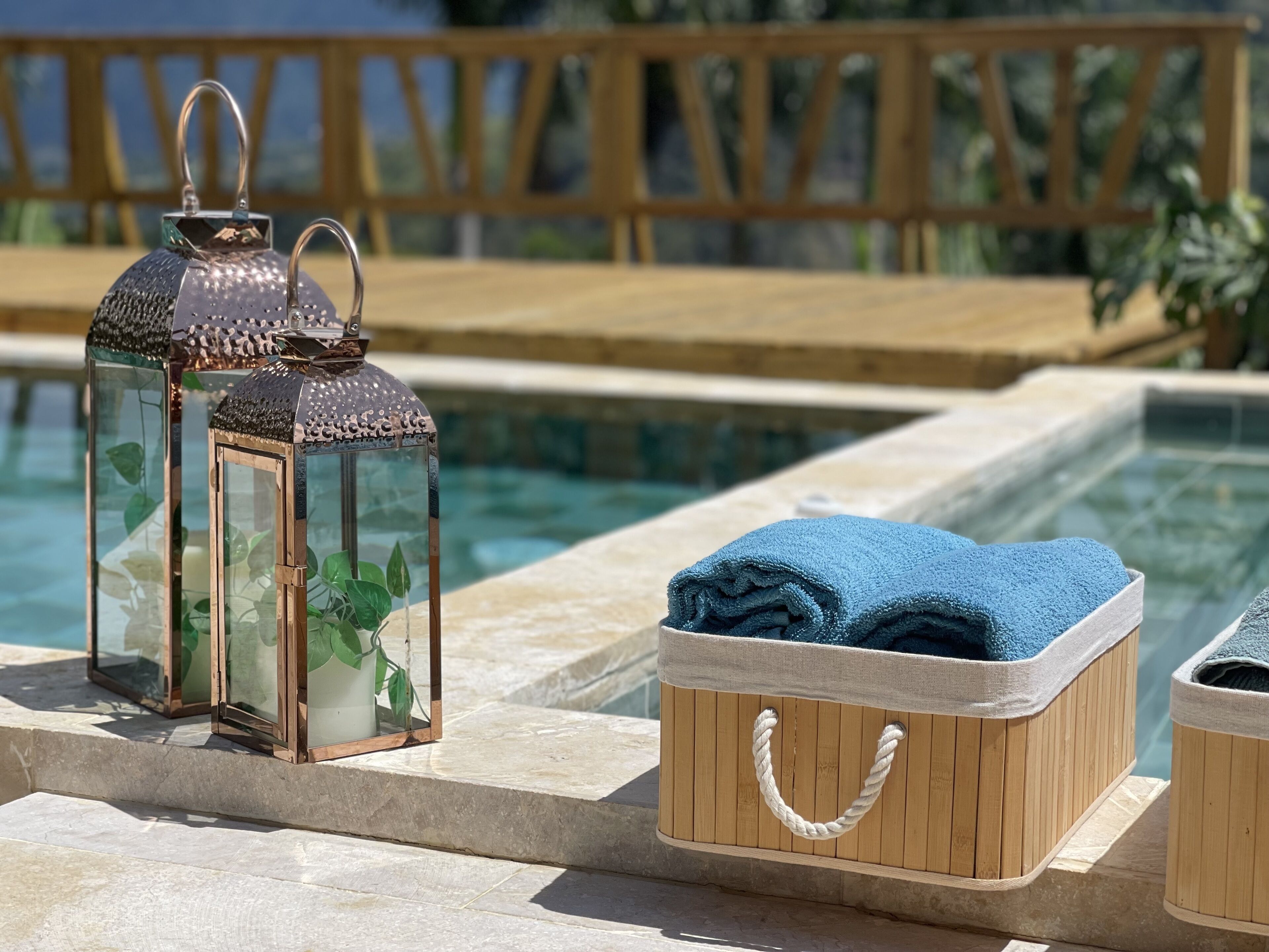 Un espace aménagé au bord de la piscine, avec une terrasse en bois, une lanterne en verre abritant une plante, et une boîte en bois contenant une serviette.