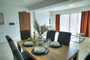 A dining area with a wooden table set for four, surrounded by grey chairs. There is a vase with dried plants on the table, and a bottle of wine and glasses are also visible.