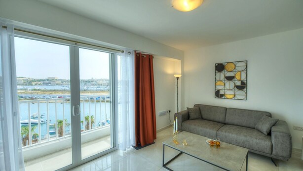 Un salon moderne avec un canapé, une table basse et une vue sur une marina, visible à travers de grandes fenêtres.