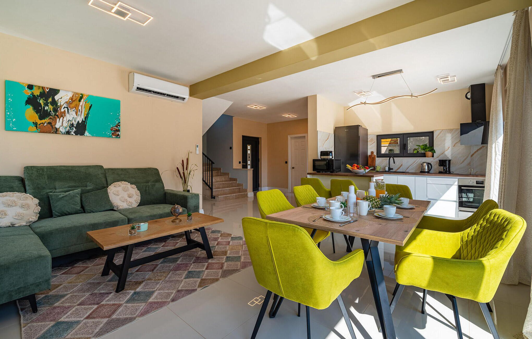 Una sala de estar con un sofá verde, una mesa de centro de madera y sillas de color amarillo brillante. Se ve una cocina al fondo, con un microondas y un televisor.
